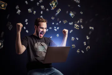 A man celebrating a winning moment on his laptop while gaming symbols float around him, capturing the excitement of 77ABC games.